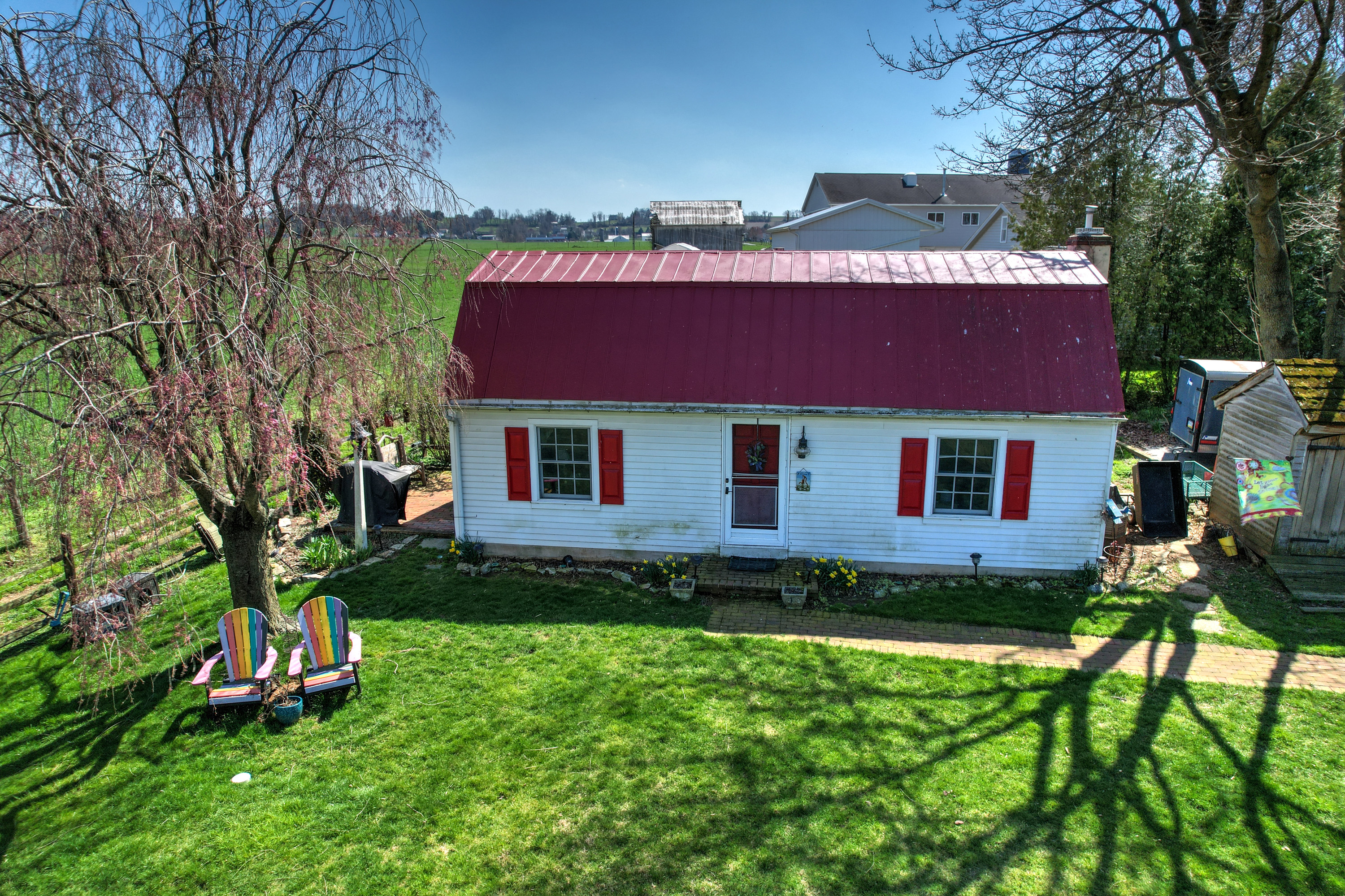 A quaint white cottage with a vibrant red roof and matching window trim, featuring a well-maintained green lawn and two colorful Adirondack chairs.