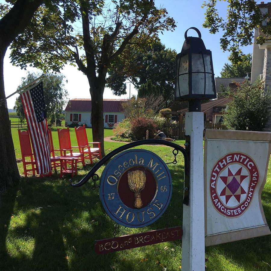 A sign for Osceola Mill House Bed and Breakfast, with American flags and red rocking chairs in the background.