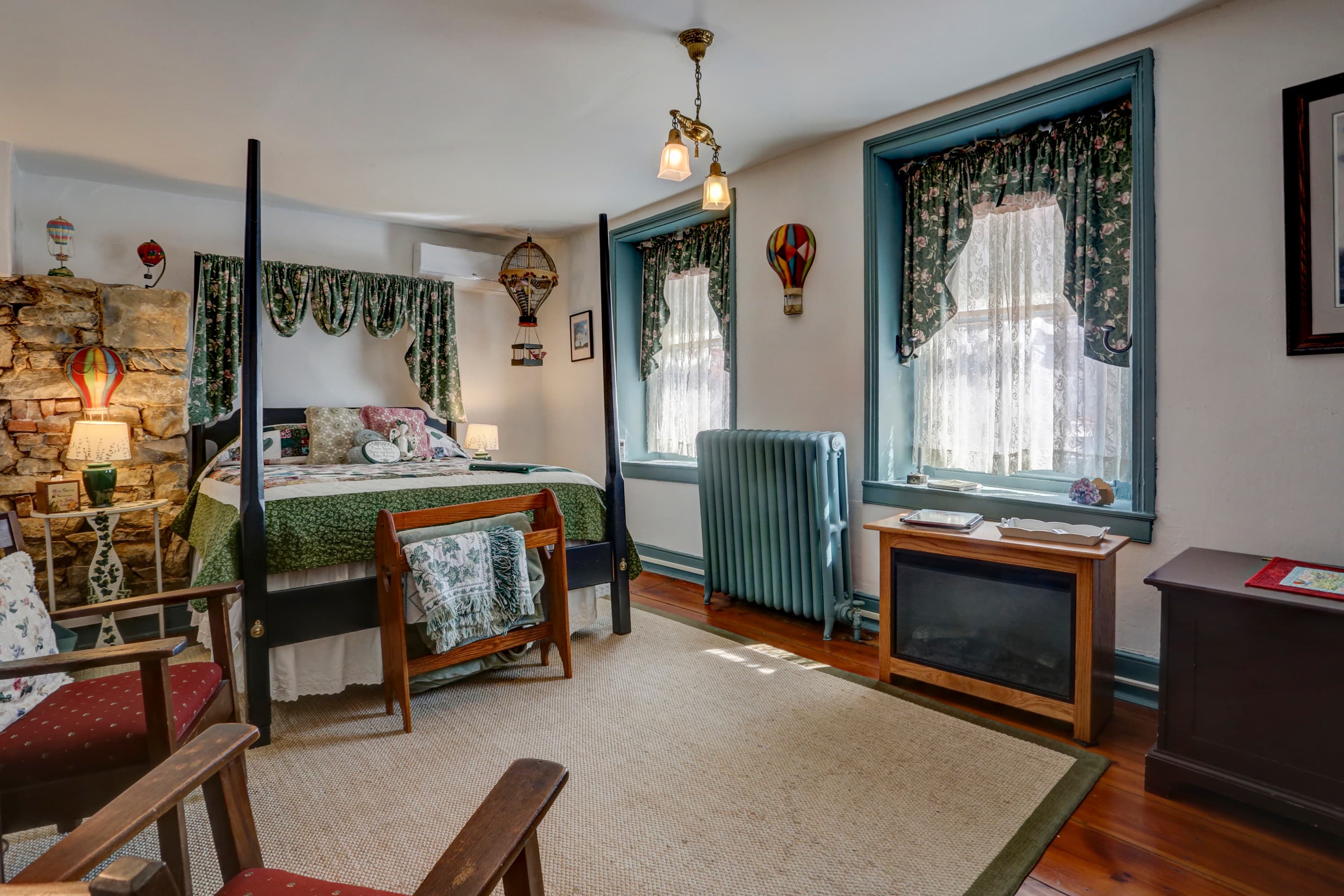 Bedroom with four-poster bed with a green comforter, two armchairs, and two windows with blue trim and floral curtains.