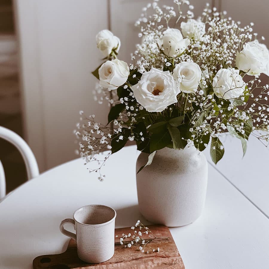 A bouquet of white roses and baby's breath in a ceramic vase sits on a table beside a small cup and a wooden board.