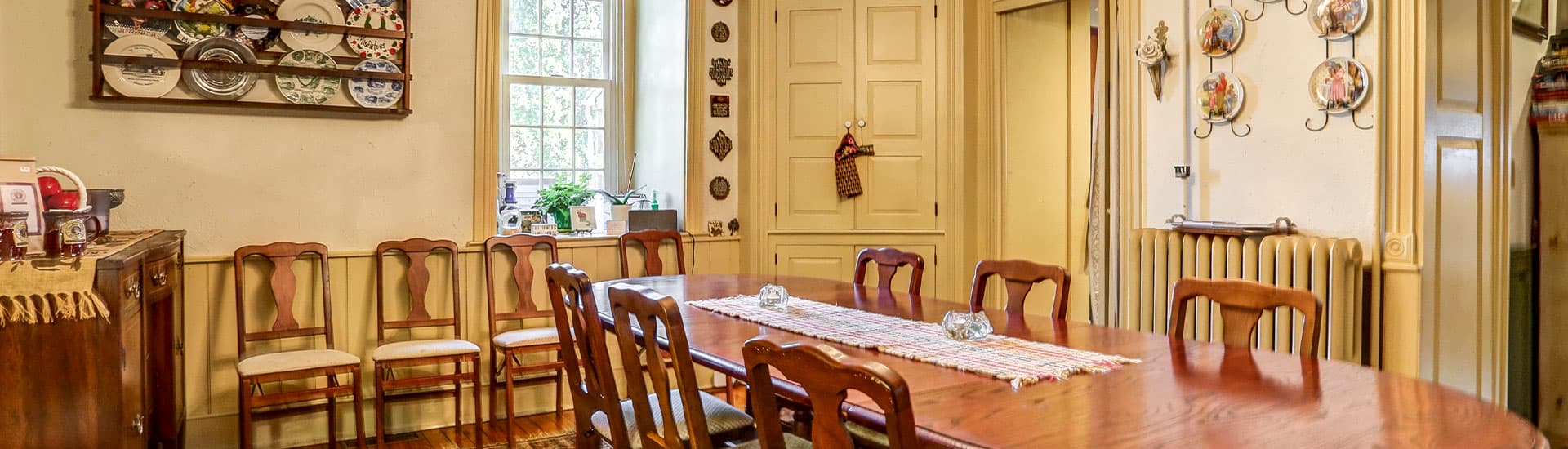 A warm, inviting kitchen featuring a large wooden dining table, wooden chairs, and colorful decorative plates on the walls.