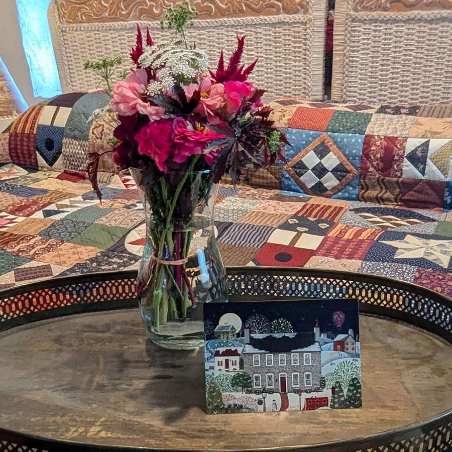 A vibrant bouquet of flowers sits in a vase on a wooden table, beside a decorative card, with a quilted bedspread in the background.