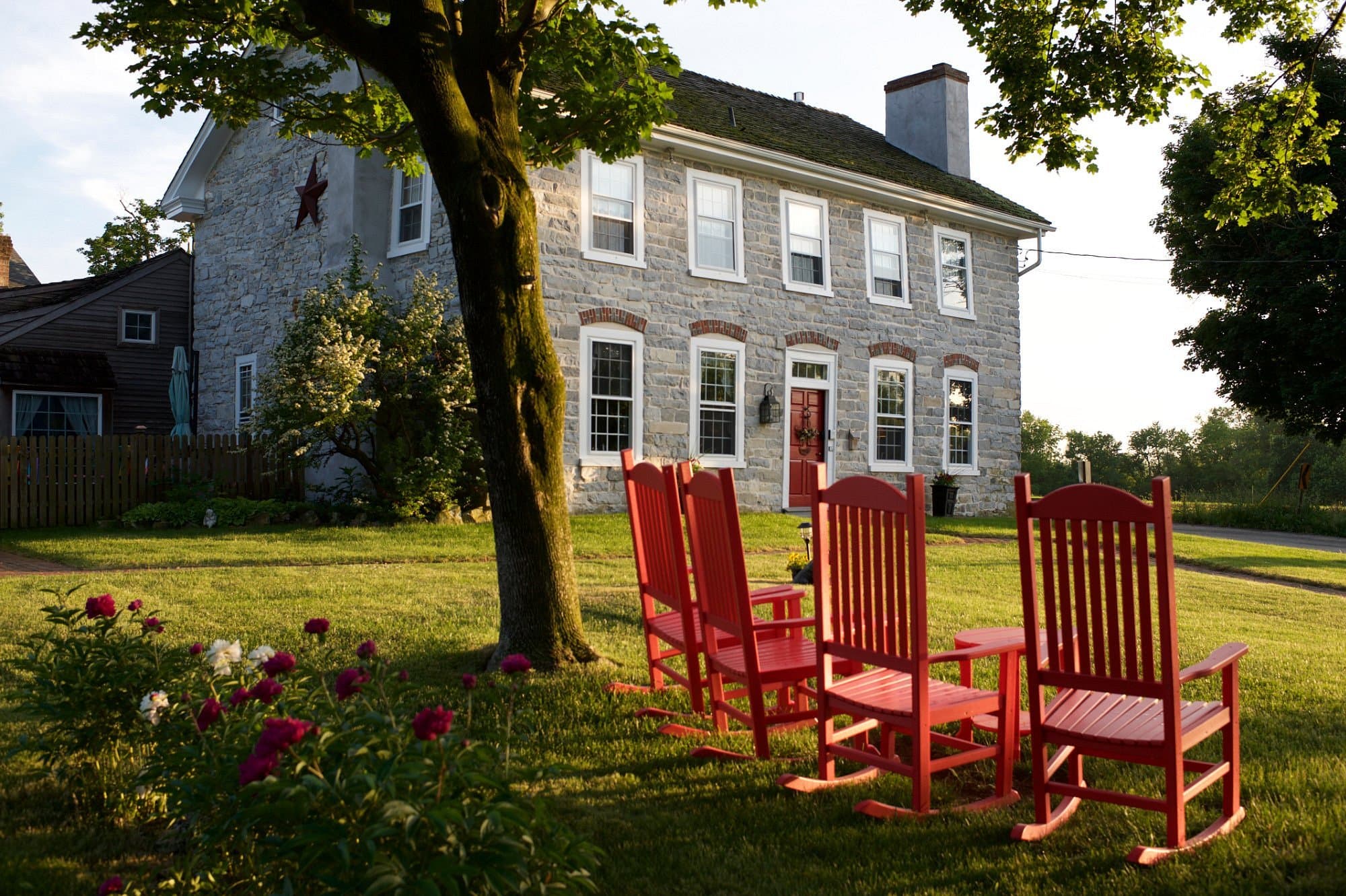 A stone house with large windows is surrounded by a grassy yard featuring four red rocking chairs and blooming flowers. A stone house with large windows is surrounded by a grassy yard featuring four red rocking chairs and blooming flowers.