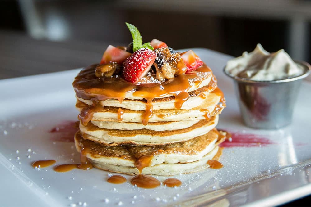 A tall stack of pancakes drizzled with caramel sauce, topped with strawberries and a side of whipped cream.