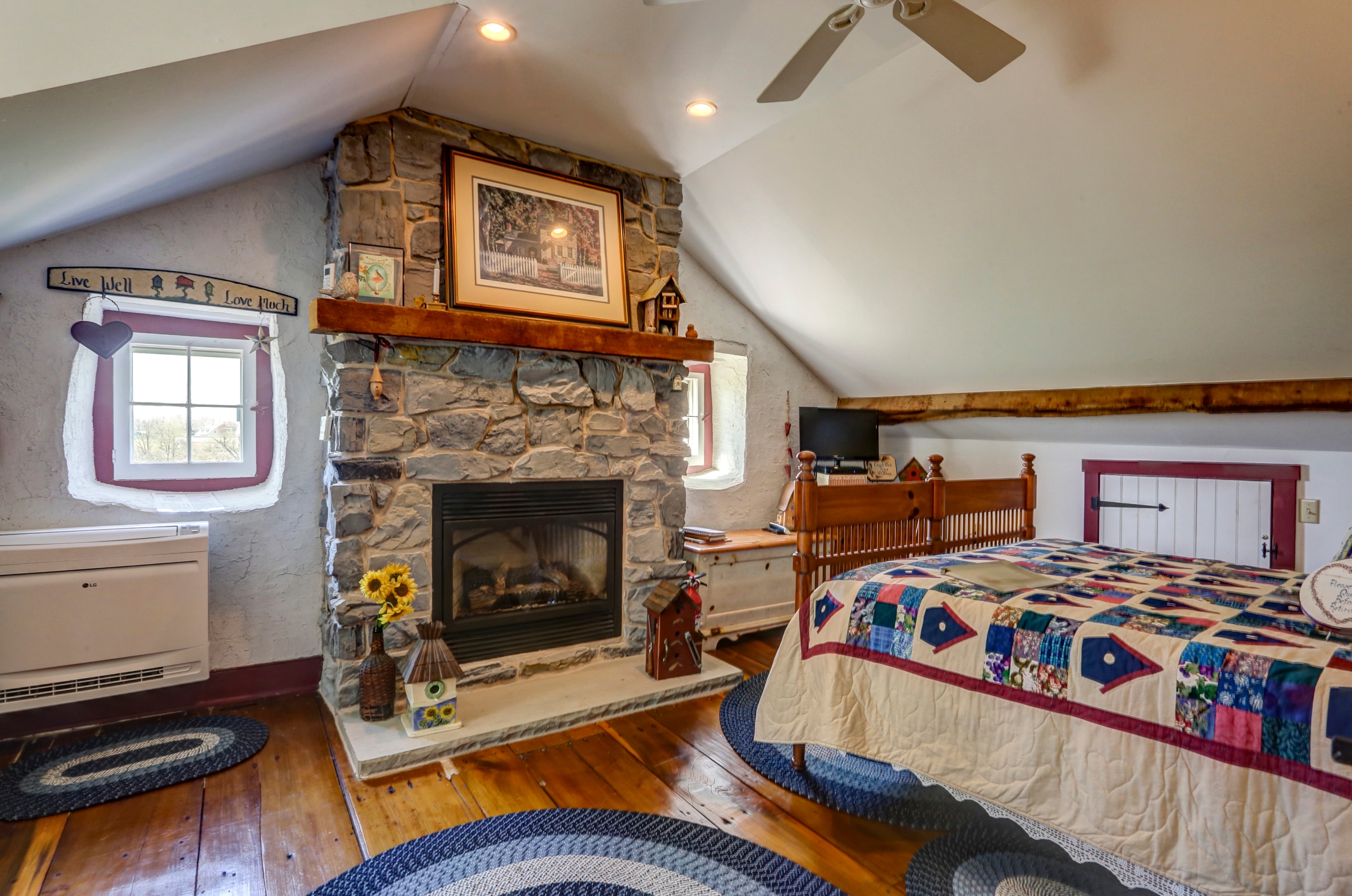 Bedroom with a focus on a stone fireplace, a bed with a colorful quilt, and hardwood floors with braided oval rugs.