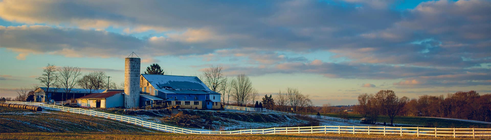 A blue farmhouse and silo surrounded by winter scenery and a white fence under a cloudy sky.