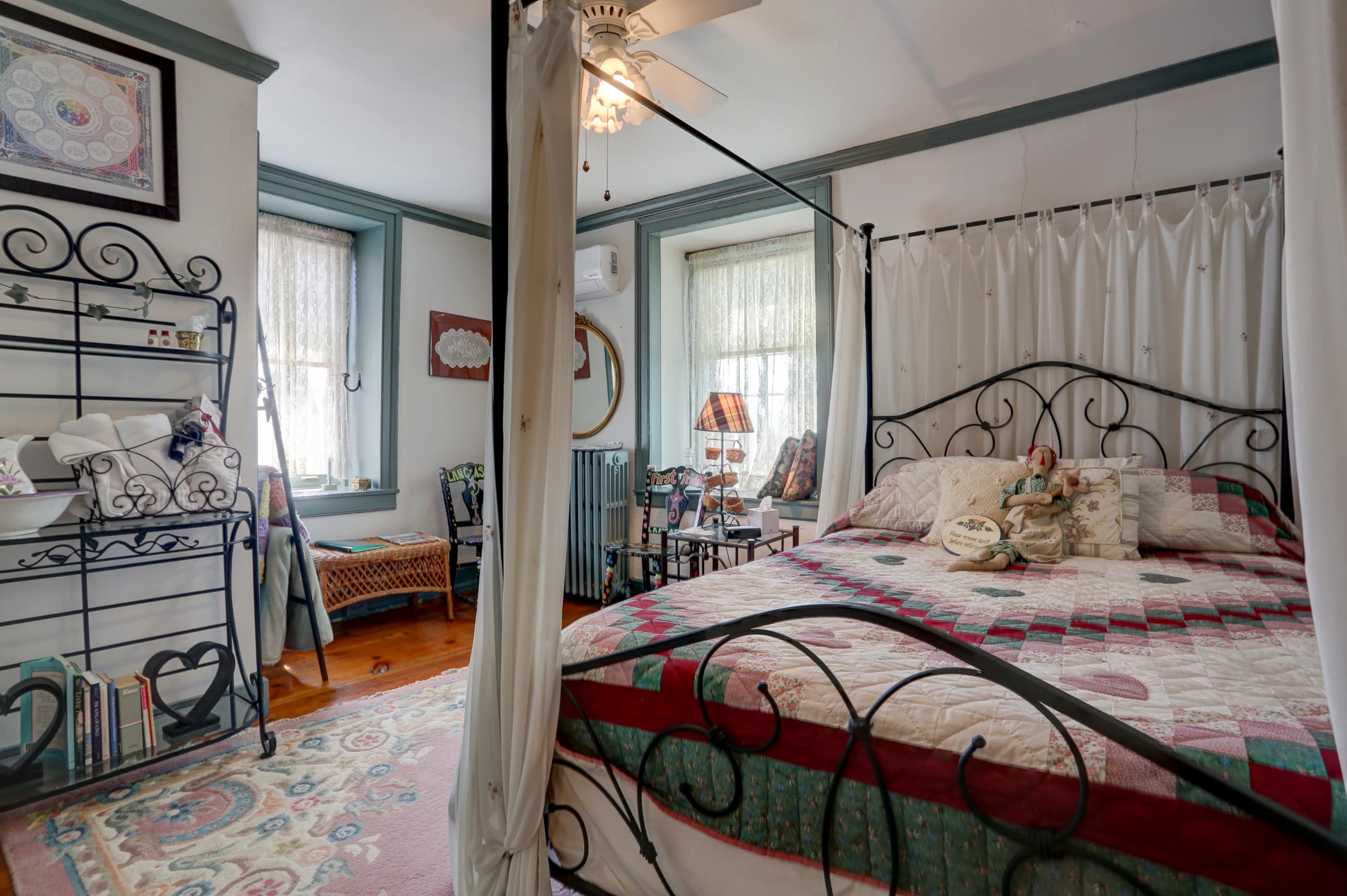 Bedroom with a patterned quilt on a decorative iron bed, two windows with white curtains, and a dresser with a mirror.